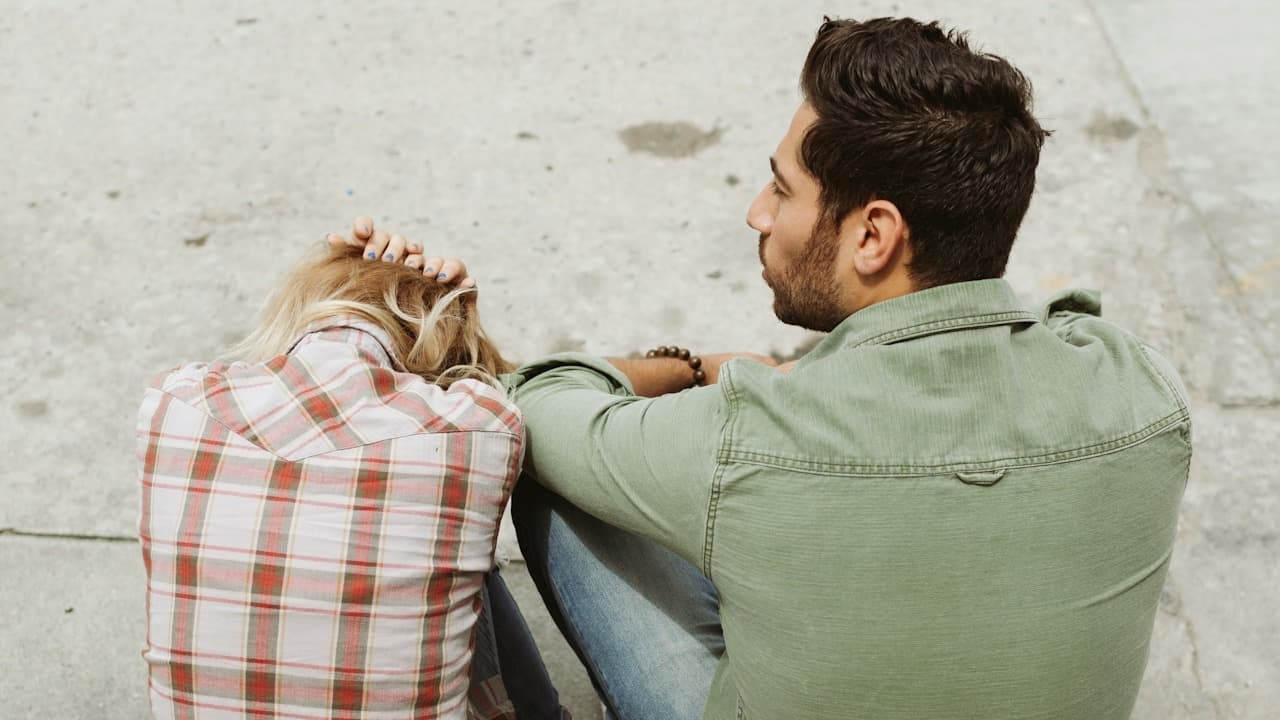 A man and a women sitting on the ground, the woman is holding her hands in her head while the man is looking into the distance